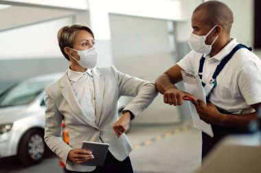 Female manager and African American mechanic elbow bumping while wearing protective face masks in auto repair shop. 
