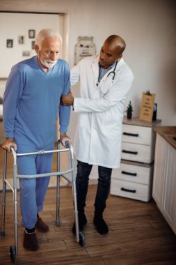 Black doctor assisting elderly man in walking with mobility walker at nursing home. 