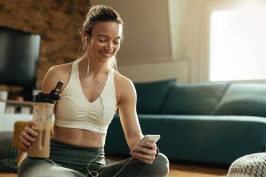 Mutlu sporcu kadın evde çalıştıktan sonra protein içeceği içerken akıllı telefon kullanıyor.. 