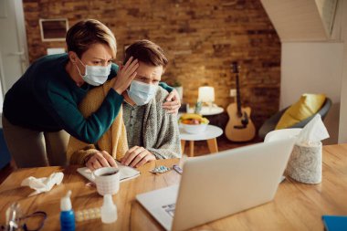 Man with fever and his worried wife looking for advice on the internet while using laptop at home during coronavirus pandemic. 