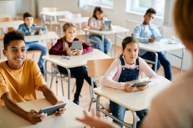 African American elementary student and her classmates e-learning on digital tablets while teacher is giving them lecture in the classroom. 