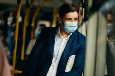 Businessman wearing protective face mask and looking through the window while traveling by public bus during coronavirus pandemic. 