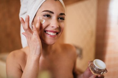 Beautiful woman wrapped in towel applying face cream in the bathroom. 