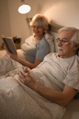 Üst düzey bir adam, kitap okuyan karısıyla yatak odasında dinlenirken cep telefonu kullanıyor.. 
