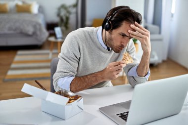 Young distraught businessman eating while reading problematic e-mail on a computer at home. 