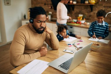 Distraught African American father working on laptop at home. His family is in the background. 