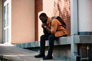 Young distraught African American student sitting at campus and thinking of something.