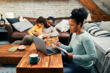 African American woman going through paperwork while working on laptop at home. Her husband and daughter are coloring in the background. 