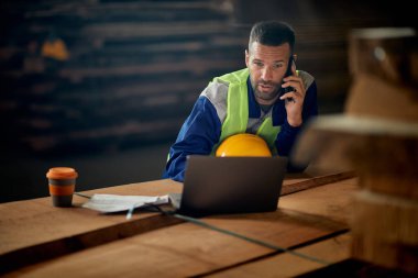 Male worker using laptop and talking on the phone while working and wood distribution warehouse. 