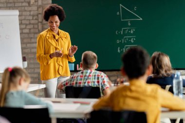 Happy black teacher talking to elementary students while giving a lesson in the classroom. 