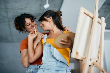 Young happy woman painting on canvas and having fun with her girlfriend at home. 