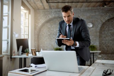 Male entrepreneur reading an e-mail on a computer and taking notes while working in the office. 