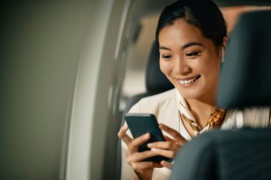 Happy Asian female CEO text messaging on mobile phone sitting on the backseat of a car.