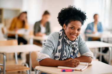 Happy African American mid adult student writing while having lecture in the classroom and looking at camera. 