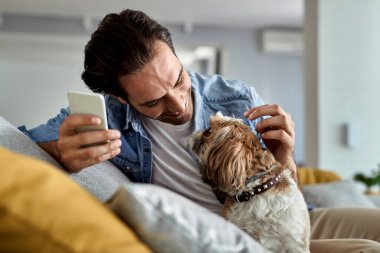 Young happy man using mobile phone and having fun with his dog in the living room. 