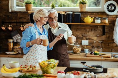 Happy senior couple surfing the net on touchpad while drinking coffee in the kitchen. 