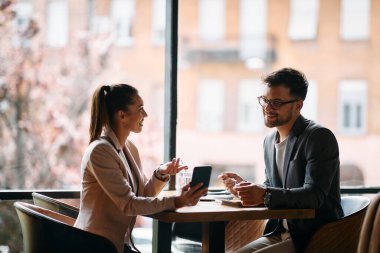 Young businesswoman showing something on smart phone to her colleague while being in a cafe. Focus is on businessman.