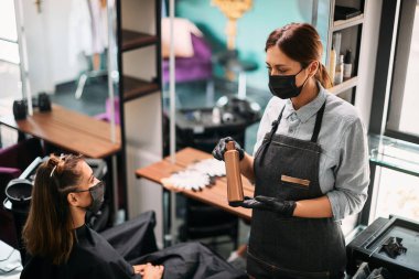 Female hairstylist and her client communicating about hair product during appointment at hair salon. They are wearing protective face masks due to coronavirus pandemic.