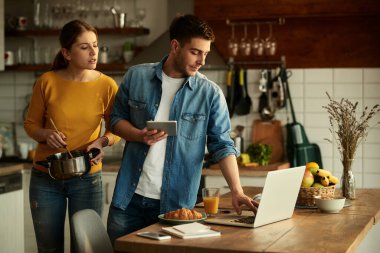 Young man working on touchpad and laptop while his wife is cooking at home.