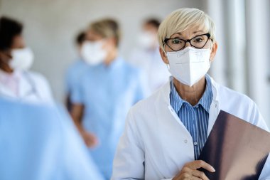 Portrait of mature female doctor wearing protective face mask while standing in hallway at medical clinic and looking at camera. 