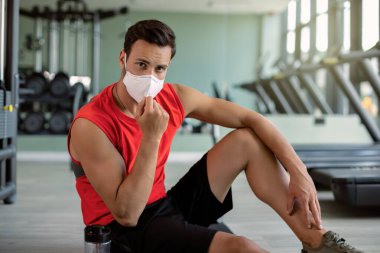 Athletic man sitting on the floor at health club while wearing protective face mask during coronavirus epidemic. clipart