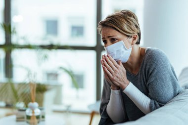 Worried woman praying with hands clasped while wearing protective face mask and feeling sad during coronavirus pandemic. 
