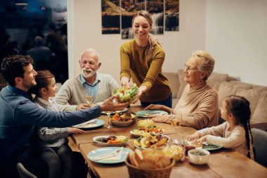 Happy extended family having a meal together at dining table. Focus is on young woman bringing salad at the table. 