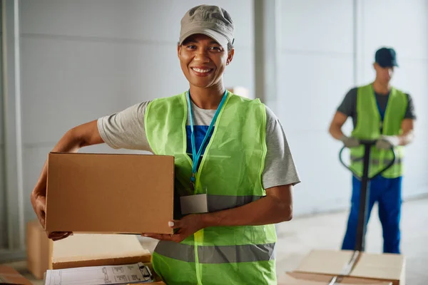 Happy African American female worker organizing cardboard boxes for shipping while working at distribution warehouse and looking at camera.