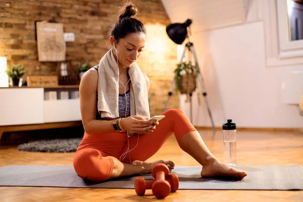 Athletic woman text messaging on mobile phone while sitting on exercise mat in the living room.