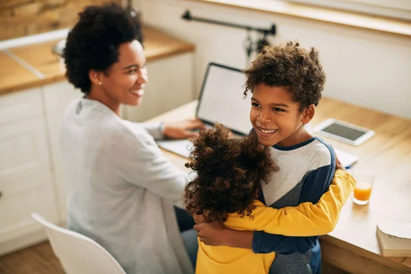 Happy black sister and brother embracing while their mother is working on laptop at home.