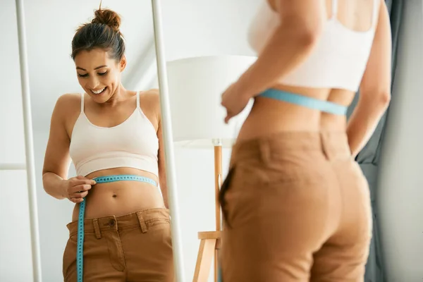 Reflection in a mirror of young woman feeling satisfied while measuring her waist.