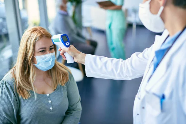 Doktor bekleme odasında sıcaklığını kızılötesi termometreyle ölçerken yüz maskesi takan kadın hasta..