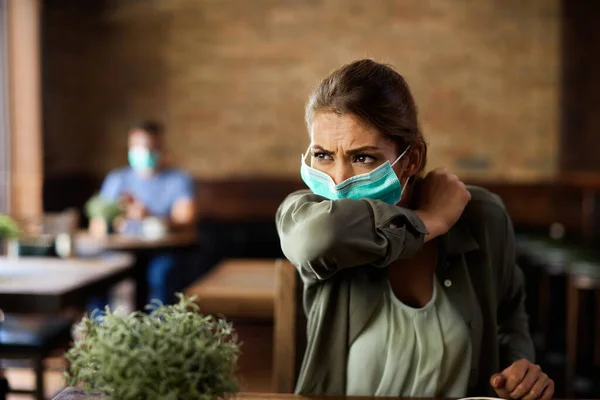 Young woman coughing into elbow while wearing protective face mask in a cafe.