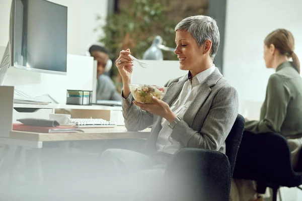 Happy female entrepreneur enjoying in lunch break and eating salad at work.