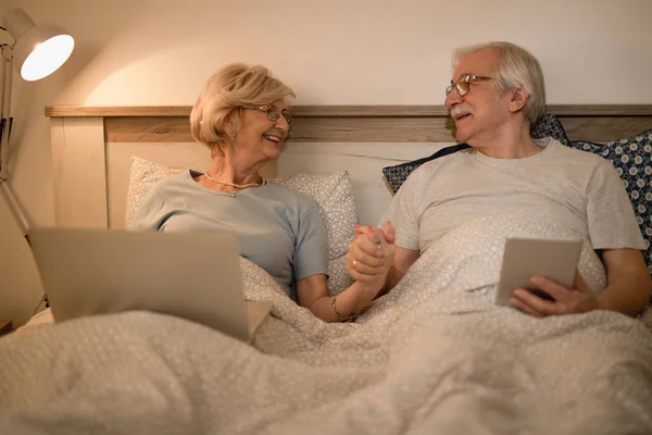 Happy senior couple talking while holding hands and using wireless technology in bed at night.
