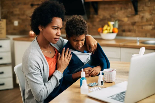 Worried black mother having online appointment with a doctor while measuring son\'s temperature at home.