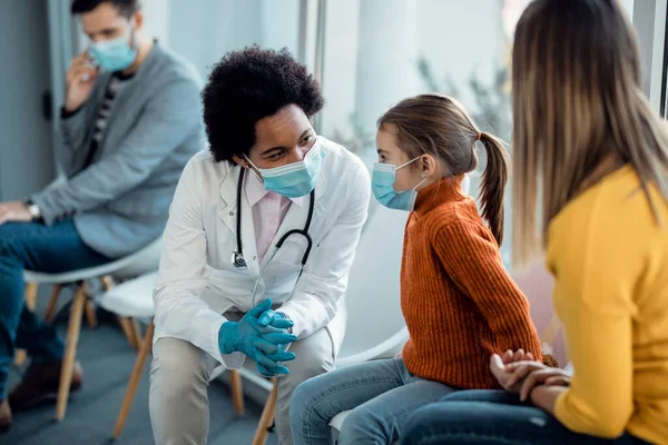 Black female doctor talking to a small girl who is sitting with her mother at hospital waiting room and wearing protective face mask due to coronavirus pandemic.