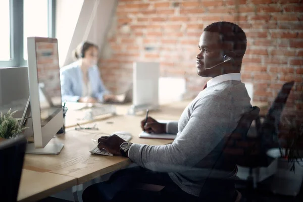 African American businessman taking notes while working on computer at corporate office. The view is through the glass.