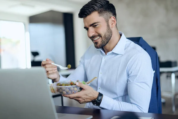 Young entrepreneur eating healthy food while using computer during lunch break in the office.
