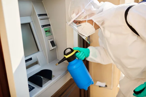 Sanitation worker in hazmat suit spraying ATM with disinfectant during coronavirus pandemic.