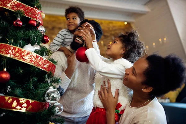 Happy black family having fun while decorating Christmas tree in the living room. Focus is on girl.