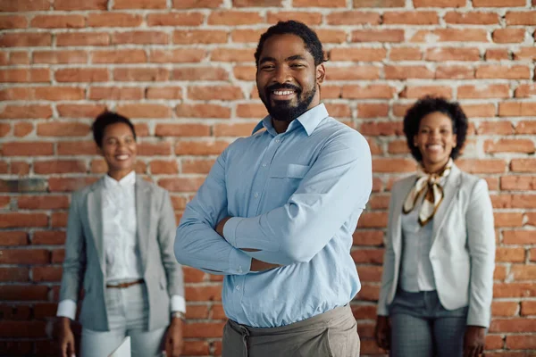 Confident black businessman standing with arms crossed and looking at camera. His female colleagues are in the background.