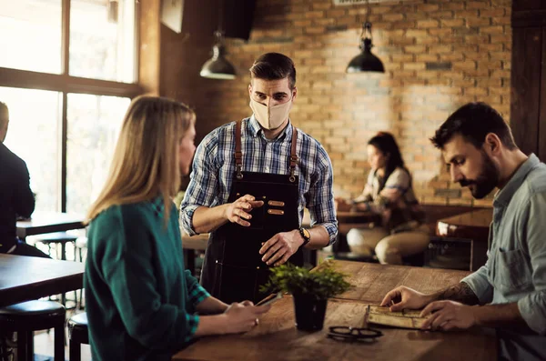 Young waiter wearing face mask while talking to guests in a pub.