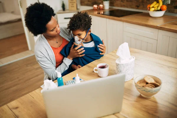 Black mother using nebulizer during inhaling therapy of her ill son at home.