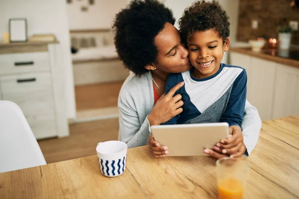 Affectionate black mother kissing her son who is sitting on her lap and surfing the net on touchpad at home.