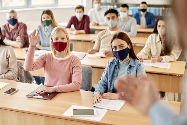 Large group of college students wearing face masks while attending lecture during coronavirus pandemic. Focus is on happy student raising her hand to answer teacher's question.