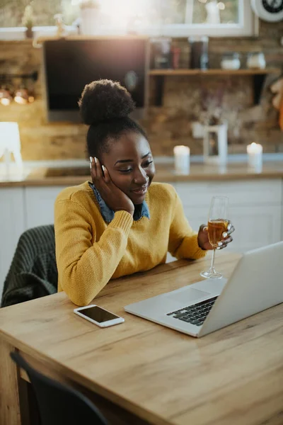 Young happy black woman using computer and making video call while drinking Champagne at home.