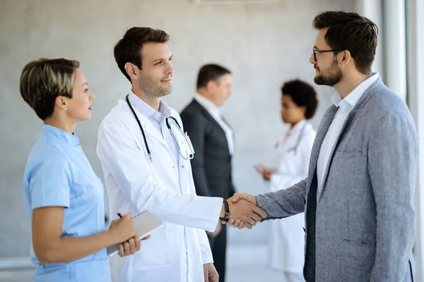 Businessman greeting healthcare workers and handshaking with a doctor in hallway at medical clinic. Focus is on male doctor.