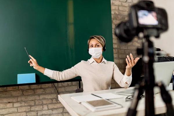 Teacher with face mask live streaming while giving lesson from the classroom during coronavirus pandemic.