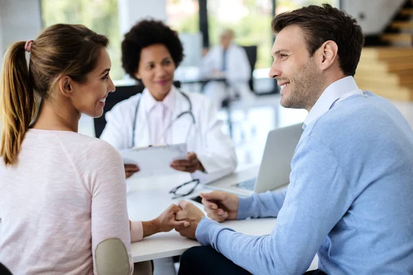 Smiling couple holding hands and talking while having consultations with a doctor at medical clinic. Focus is on man.